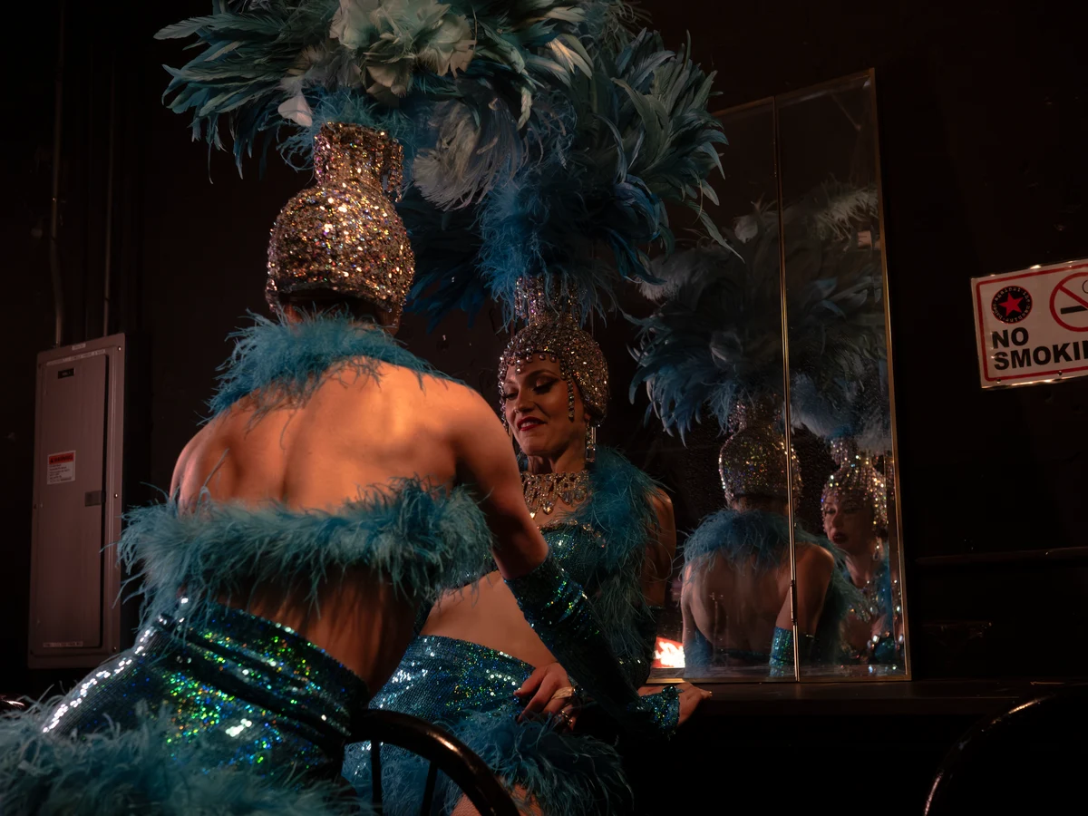 Dvě showgirls v zákulisí v kostýmech s flitry a peřím, jedna odrážející se v zrcadle (reprezentativní fotografie, nikoli vlastní soubor tohoto podniku)
